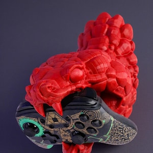 May include: A red, textured snake head with open jaws holding a black and gold video game controller. The controller has green accents and a patterned design. The snake's tongue is also red.