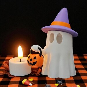 May include: A white ghost figure wearing a purple and orange witch hat, holding an orange jack-o'-lantern bucket. A lit white candle and Halloween candies are on a black and orange checkered surface. Halloween-themed decor.