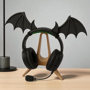 May include: Black headphones with a bat wing and horn design, displayed on a wooden stand. The headphones have a built-in microphone and are on a light-coloured wooden surface.