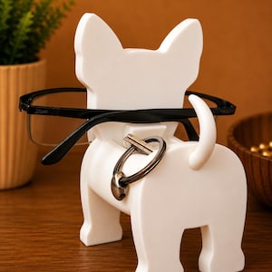 May include: A white bulldog-shaped eyeglasses and key holder. Black eyeglasses rest on the dog's head, and a key ring hangs from its back. The holder is on a wooden surface, with a plant and a wooden bowl in the background.