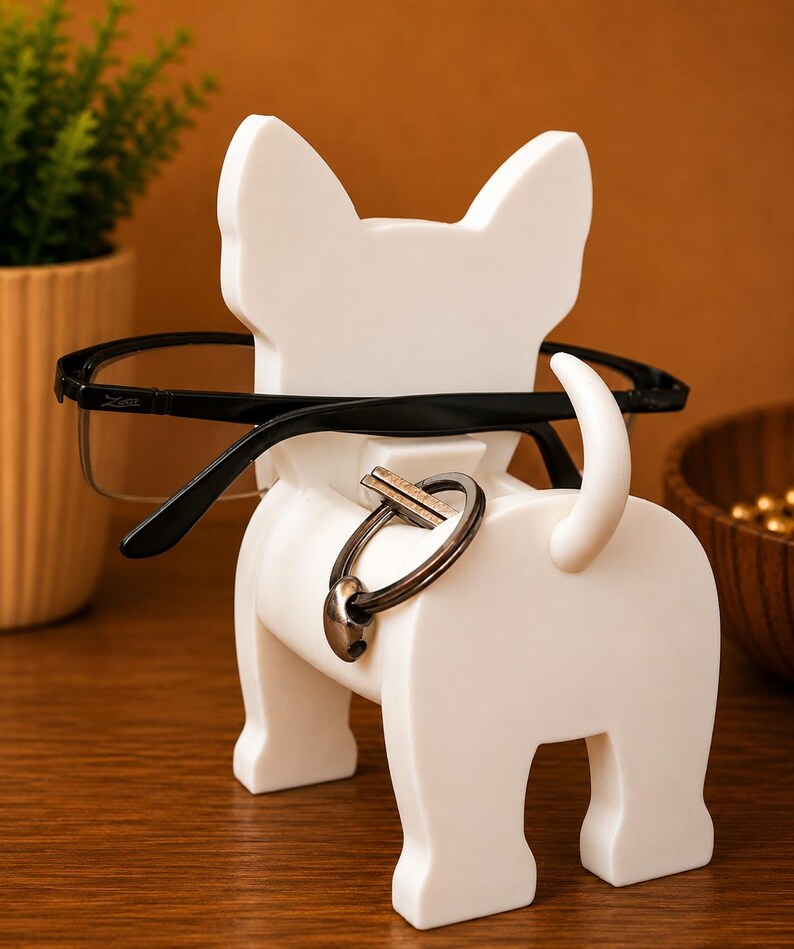 May include: A white bulldog-shaped eyeglasses and key holder. Black eyeglasses rest on the dog's head, and a key ring hangs from its back. The holder is on a wooden surface, with a plant and a wooden bowl in the background.