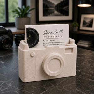 May include: A beige camera-shaped business card holder with a stack of white cards. The cards display the name "Jane Smith Photography", contact information, and a lens graphic. The holder has a textured surface and a lens-shaped front.
