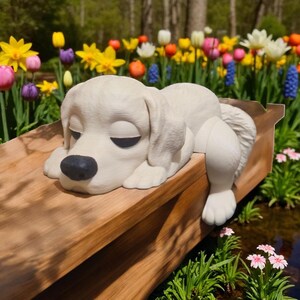 May include: A white, sleeping dog statue with a black nose and closed eyes rests on a wooden surface. The statue is surrounded by colorful flowers, including tulips and daffodils, creating a serene garden scene.
