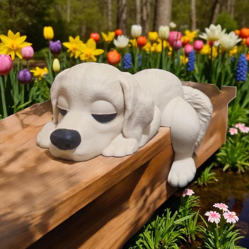 May include: A white, sleeping dog statue with a black nose and closed eyes rests on a wooden surface. The statue is surrounded by colorful flowers, including tulips and daffodils, creating a serene garden scene.