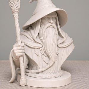 May include: A detailed white bust of a wizard with a long beard, a pointed hat, and a staff. The wizard's face has a stern expression. The staff has a crystal-like top. The bust is on a round base, and the wizard is wearing a robe.