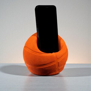 May include: An orange basketball-shaped phone holder with a black smartphone inside. The holder has a textured surface with the typical lines of a basketball. The phone is upright, and the holder is on a light-coloured surface.