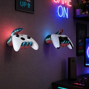 May include: Two wall-mounted controller holders shaped like metallic monster hands with sharp claws. Each hand is painted in a gradient of blue and silver and securely grips a white wireless gaming controller against a dark purple wall.