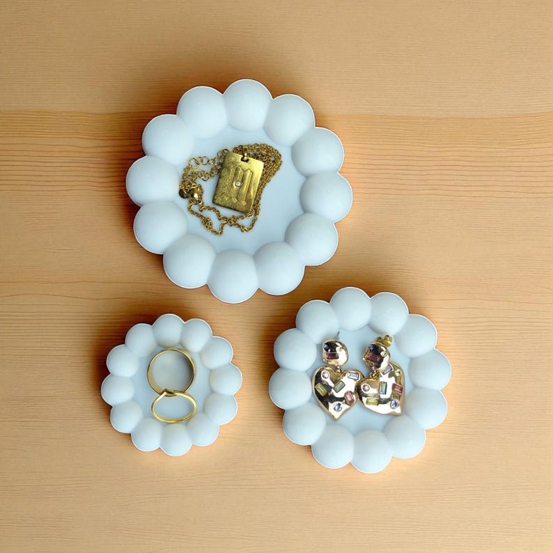 May include: Three white, cloud-shaped trinket dishes with gold jewelry. One dish holds a gold necklace with a rectangular pendant. Another holds two gold rings. The third dish contains two heart-shaped earrings with gold and jewel accents.