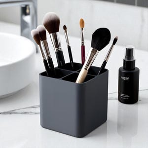 May include: A dark gray makeup brush holder with multiple compartments, filled with various makeup brushes. A black spray bottle with white text is to the right. The brushes have black handles and soft bristles.