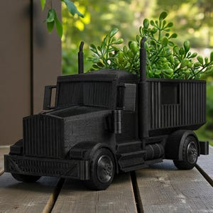 K&ouml;nnte beinhalten: Ein schwarzer dekorativer Pflanzk&uuml;bel in Form eines Sattelzugs. Die Ladefl&auml;che des Lastwagens ist mit gr&uuml;nen Pflanzen gef&uuml;llt. Der Pflanzk&uuml;bel besteht aus einem strukturierten Material und steht auf einer Holzoberfl&auml;che.