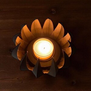 May include: A lit candle in a decorative, flower-shaped holder. The holder features orange petals with black accents, creating a layered effect. The candle is centered within the holder, casting a warm glow. The candle holder is on a dark wooden surface.
