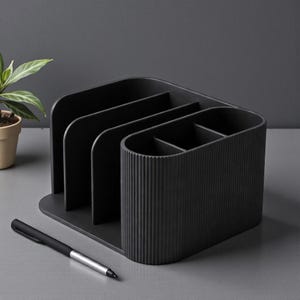 May include: A black desk organizer with multiple compartments for storing office supplies. The organizer has a ribbed texture on one side and a pen is placed in front of it. A small potted plant is visible in the background.