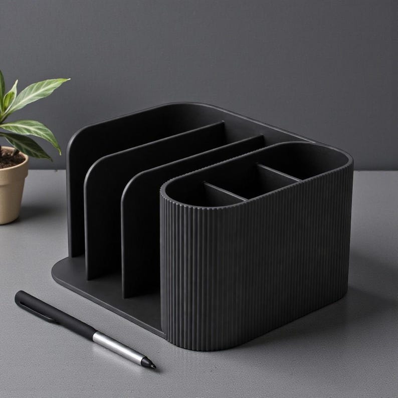 May include: A black desk organizer with multiple compartments for storing office supplies. The organizer has a ribbed texture on one side and a pen is placed in front of it. A small potted plant is visible in the background.
