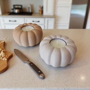 Puede incluir: Dos candelabros con forma de calabaza, uno beige y otro blanco, cada uno con una vela blanca encendida. Un cuchillo de acero inoxidable con mango texturizado est&aacute; cerca. Sobre una encimera clara.
