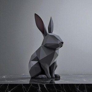 May include: A geometric, dark gray rabbit sculpture with a faceted design. The rabbit has long ears with pink interiors and a red nose. It sits on a black and white marble surface against a gray background. The sculpture is a modern home decor item.