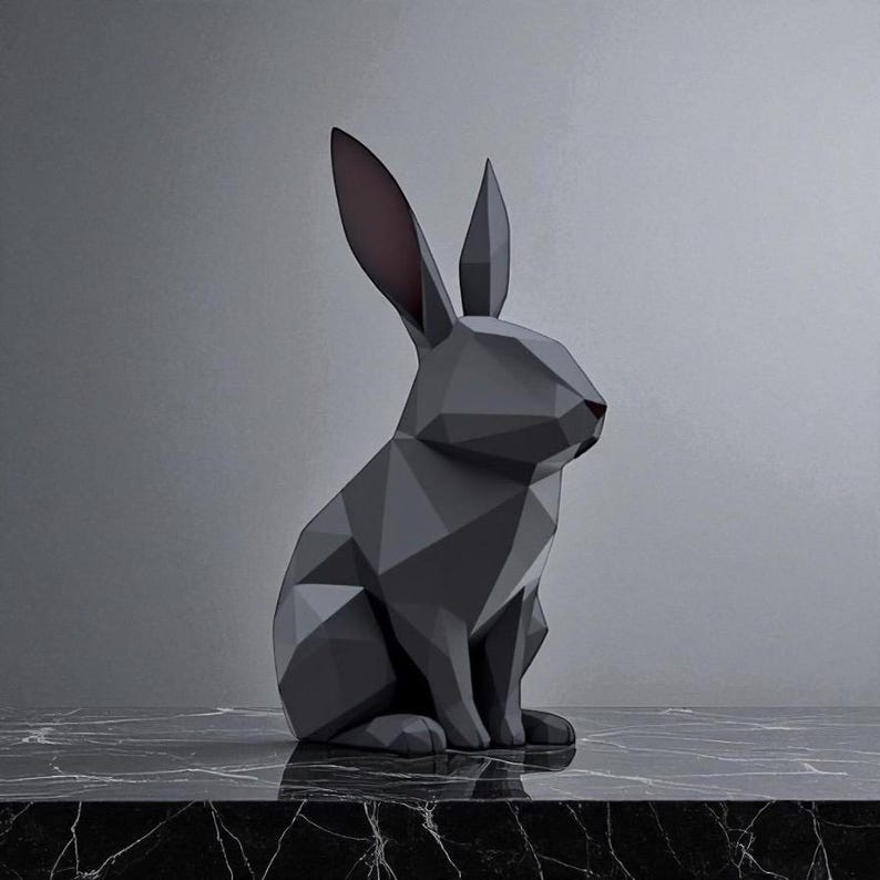 May include: A geometric, dark gray rabbit sculpture with a faceted design. The rabbit has long ears with pink interiors and a red nose. It sits on a black and white marble surface against a gray background. The sculpture is a modern home decor item.