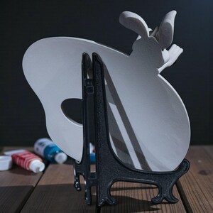 May include: A light grey ceramic palette-shaped plate with a bunny design, displayed on a black easel. The plate has a hole for the thumb and is set against a dark background. Paint tubes are visible in the background.