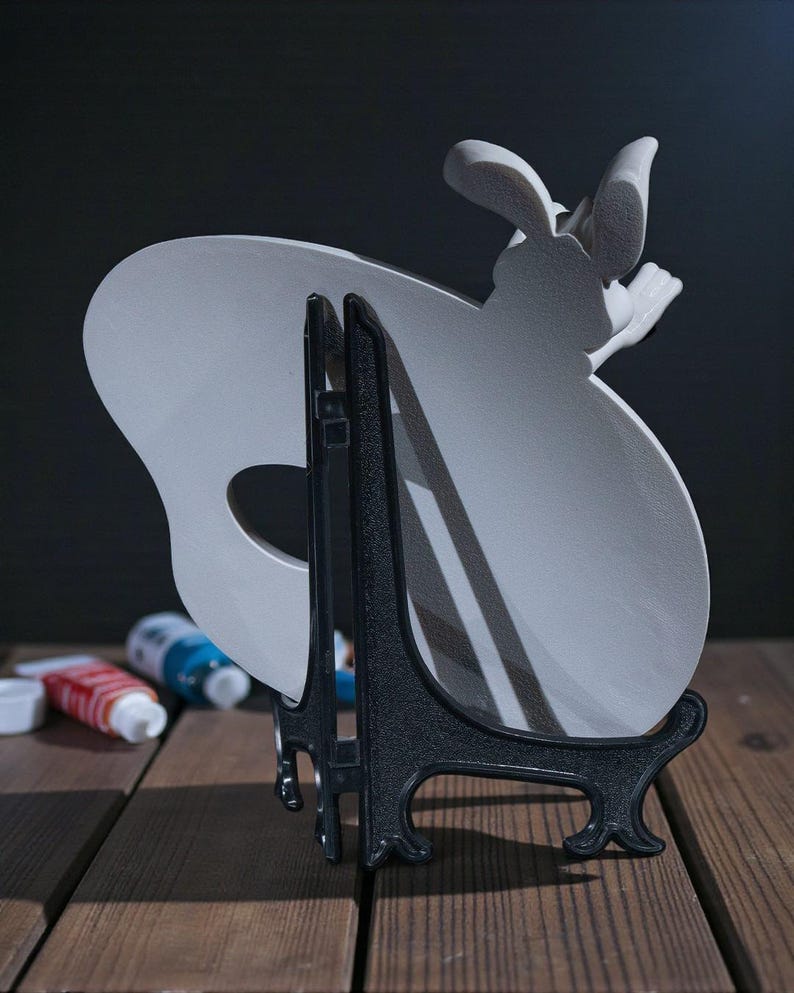 May include: A light grey ceramic palette-shaped plate with a bunny design, displayed on a black easel. The plate has a hole for the thumb and is set against a dark background. Paint tubes are visible in the background.