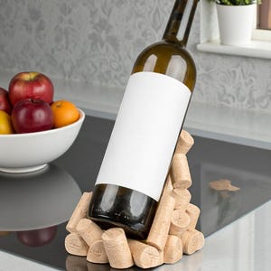 Puede incluir: Una botella de vino con una etiqueta en blanco se muestra en un soporte para botellas de vino de corcho. El soporte est&aacute; hecho de corchos apilados. Un cuenco de fruta est&aacute; en el fondo.