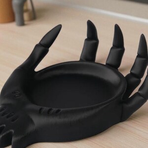 May include: A black hand-shaped dish with a shallow bowl and extended fingers. The dish is made of a solid material and sits on a light-colored surface. The fingers have pointed tips.