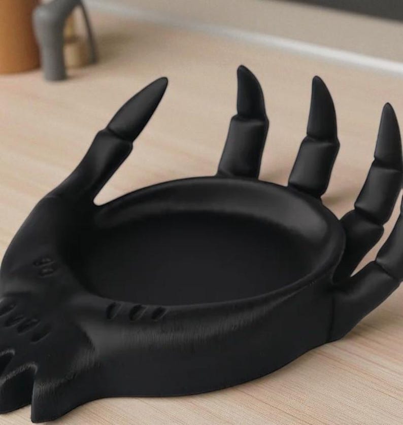 May include: A black hand-shaped dish with a shallow bowl and extended fingers. The dish is made of a solid material and sits on a light-colored surface. The fingers have pointed tips.