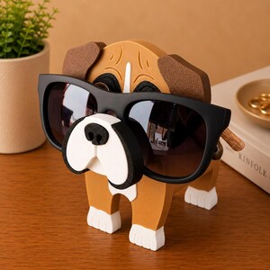Peut inclure: Un porte-lunettes en forme de chien en bois avec des lunettes de soleil noires. Le support est marron, blanc et noir, avec les lunettes de soleil pos&eacute;es sur le nez du chien. Le chien a des pattes blanches et un visage blanc et noir.