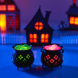May include: Two black cauldrons with glowing green and pink liquid inside. The cauldrons are sitting on a blue surface in front of a miniature Halloween village.