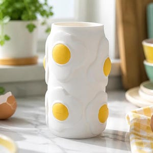 May include: A white ceramic vase with a repeating pattern of fried eggs. Each egg has a yellow yolk and a white, textured egg white. The vase is cylindrical and stands on a marble countertop.