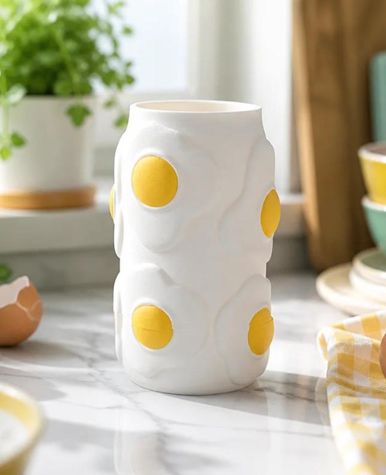 May include: A white ceramic vase with a repeating pattern of fried eggs. Each egg has a yellow yolk and a white, textured egg white. The vase is cylindrical and stands on a marble countertop.