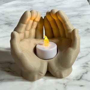 Puede incluir: Un portavelas beige en forma de manos acuna una peque&ntilde;a vela blanca con una llama amarilla parpadeante. Las manos est&aacute;n esculpidas con dedos y palmas detallados, creando una est&eacute;tica c&aacute;lida y acogedora. El portavelas est&aacute; dise&ntilde;ado para sostener una peque&ntilde;a vela de t&eacute;.