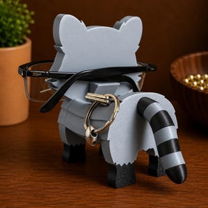 May include: A gray raccoon-shaped desk organizer holding eyeglasses and a key ring. The raccoon has a black and white striped tail and black feet. The eyeglasses are black-framed. The organizer is made of layered material.