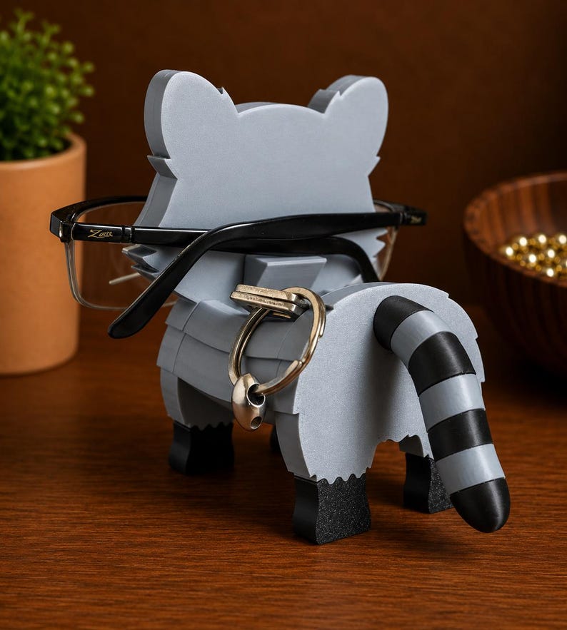 May include: A gray raccoon-shaped desk organizer holding eyeglasses and a key ring. The raccoon has a black and white striped tail and black feet. The eyeglasses are black-framed. The organizer is made of layered material.