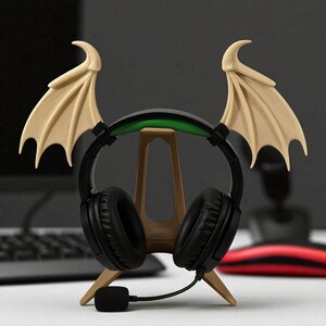 May include: A black gaming headset with a microphone rests on a tan stand with beige bat wings. The headset has a green accent on the headband. The stand is on a white surface with a computer keyboard and mouse in the background.