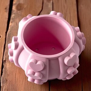 May include: A light pink planter with a round opening and heart-shaped embellishments. The planter has a textured surface and is made of a solid material. The planter is sitting on a wooden surface.
