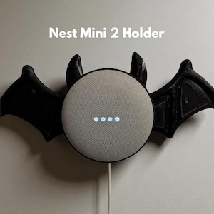 May include: A bat-shaped Nest Mini 2 holder with black wings and horns. The speaker is grey with four illuminated dots. The text "Nest Mini 2 Holder" is displayed above the speaker.