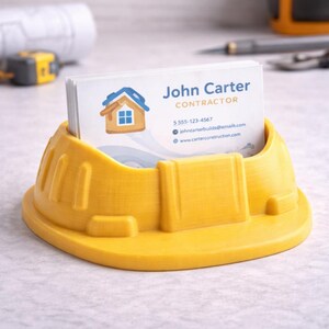 May include: A bright yellow, 3D-printed business card holder shaped like a construction hard hat. The holder contains a stack of business cards with the name "John Carter, Contractor" and contact information. The hard hat design features a curved brim and textured details.