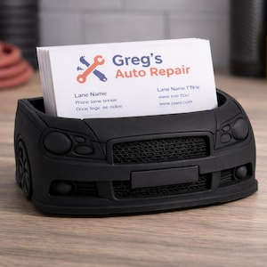 May include: A black car-shaped business card holder with a stack of white cards. The cards display the text "Greg's Auto Repair." The holder has a detailed front grille, headlights, and bumper.