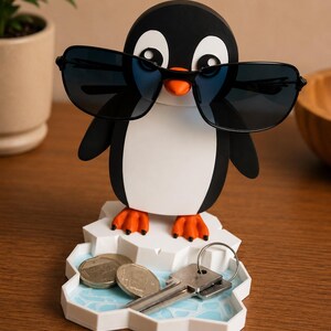 May include: A black and white penguin-shaped desk organizer with sunglasses. The penguin stands on a white iceberg-shaped tray with a light blue water design. The tray holds coins and keys.