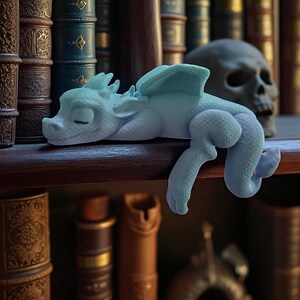 May include: A light blue dragon figurine with folded wings and a curled tail rests on a wooden shelf. The dragon is lying down with its eyes closed. A skull and antique books are in the background.