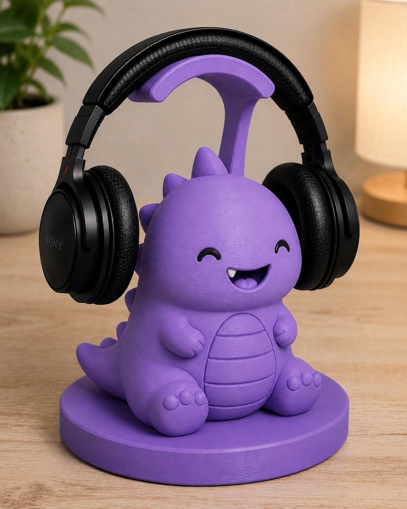 May include: A purple dinosaur-shaped headphone stand with a smiling face and black headphones resting on top. The stand is made of a solid material and sits on a round base. The headphones are black.