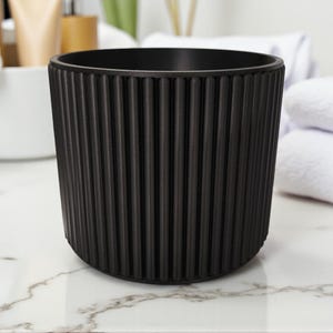 May include: A black, cylindrical container with vertical ribbed detailing. The container is made of a matte material and sits on a marble-patterned surface. The design is simple and modern, suitable for bathroom or vanity use.