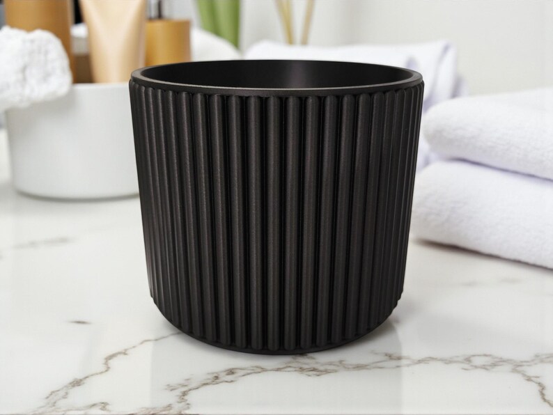 May include: A black, cylindrical container with vertical ribbed detailing. The container is made of a matte material and sits on a marble-patterned surface. The design is simple and modern, suitable for bathroom or vanity use.