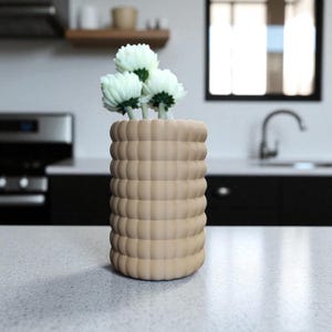 Pu&ograve; includere: Un vaso beige, strutturato, di forma cilindrica, che contiene tre fiori bianchi con centri verdi. Il vaso ha un design a coste e poggia su un piano di lavoro chiaro. Lo sfondo mostra una cucina con lavello e mobili.