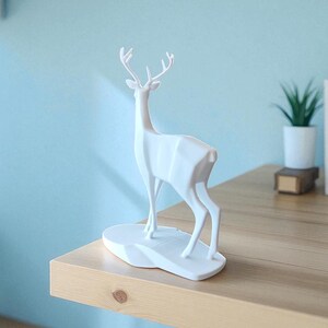 May include: A white geometric deer sculpture with antlers stands on a white base. The sculpture is on a wooden shelf, with a small potted plant and books in the background. The wall is painted a light blue.