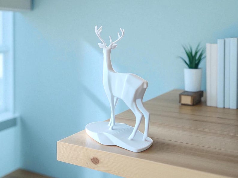 May include: A white geometric deer sculpture with antlers stands on a white base. The sculpture is on a wooden shelf, with a small potted plant and books in the background. The wall is painted a light blue.