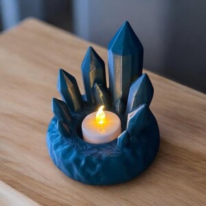 May include: A teal-colored crystal-shaped candle holder with a flickering LED candle. The holder has a textured base and is surrounded by pointed crystal formations. The candle holder is on a wooden surface.