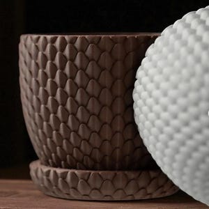 Pu&ograve; includere: Primo piano di un vaso di fiori in ceramica marrone con un motivo a scaglie in rilievo e un sottovaso abbinato. Un oggetto bianco strutturato &egrave; parzialmente visibile. Gli oggetti sono su una superficie di legno.