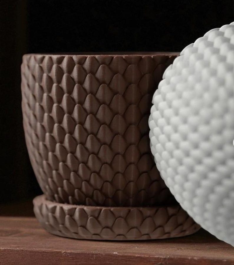 Pu&ograve; includere: Primo piano di un vaso di fiori in ceramica marrone con un motivo a scaglie in rilievo e un sottovaso abbinato. Un oggetto bianco strutturato &egrave; parzialmente visibile. Gli oggetti sono su una superficie di legno.