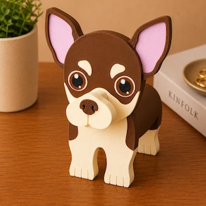 Puede incluir: Figura decorativa de chihuahua en tonos marr&oacute;n, crema y rosa. El perro tiene ojos grandes y expresivos y una nariz detallada. Las orejas son rosas por dentro. La figura est&aacute; sobre una superficie de madera.