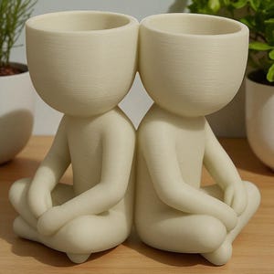 May include: Two off-white ceramic planters shaped like seated figures with their legs crossed. Each figure has a large, open head that serves as a planting pot. The figures are positioned back-to-back, creating a unique decorative piece.